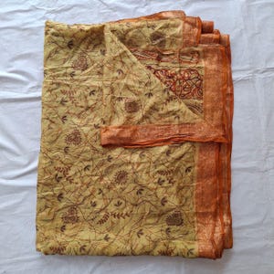 May include: A folded fabric textile featuring a light yellow base color with a dark brown linear pattern of thin, intersecting lines and small floral motifs. The edges are finished with a wide, solid orange border with a subtle texture.