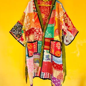 May include: A colourful patchwork kimono jacket with a variety of patterns and hues. The jacket has a green trim, two front pockets, and a matching belt. It is suspended on a yellow hanger against a yellow backdrop.