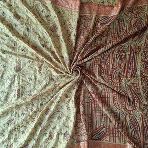 May include: A fabric textile featuring a light yellow base with a complex pattern of dark brown and reddish-brown lines, swirling paisley shapes, and small circular motifs. The material is gathered in a central spiral, creating folds.