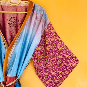May include: A vibrant kimono with a mix of patterns and colours. The kimono features a blue and pink floral design on the sleeves and a paisley pattern on the cuffs. The garment is hanging on a yellow hanger against a yellow background.