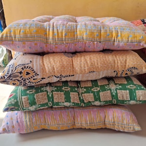 May include: A stack of four rectangular floor cushions featuring various patterns and colors. The top cushion is yellow and pink with small blue details. The second is orange with black floral motifs. The third is green with geometric squares. The bottom is pink and yellow.