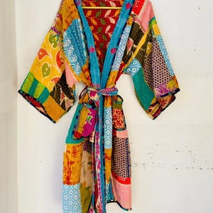 Puede incluir: Una colorida bata kimono patchwork con cinturón anudado, colgada de una percha amarilla. La bata presenta una variedad de patrones y colores, incluyendo azul, amarillo, rojo y verde. La bata está hecha de una tela ligera.