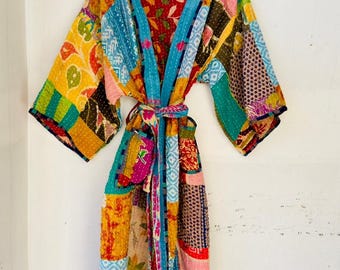 May include: A colorful patchwork kimono robe with a tie belt, hanging on a yellow hanger. The robe features a variety of patterns and colors, including blue, yellow, red, and green. The robe is made of a lightweight fabric.