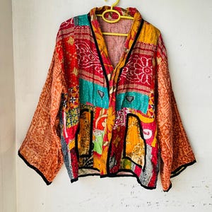 May include: A patchwork kimono-style jacket with a vibrant mix of colors, including red, orange, teal, and yellow. The jacket features a V-neck and long sleeves with black trim. The fabric appears to be quilted.