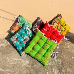 May include: A collection of colorful square seat cushions with various patterns and colors, including green, red, yellow, and teal. Each cushion has a quilted design and white tie closures. The cushions are arranged on a concrete surface.
