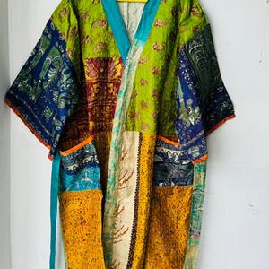 May include: A patchwork kimono-style robe in vibrant colours. The robe features a mix of patterns and textures, including floral and paisley designs. The colour palette includes green, blue, yellow, and orange. It has a teal collar and belt.