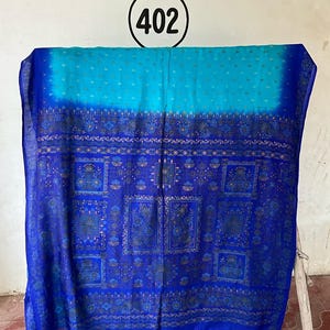 May include: A rectangular textile featuring a gradient from light blue to deep royal blue. The fabric displays a repeating pattern of dark blue floral squares and intricate geometric borders. A circular tag with the number 402 is visible above.