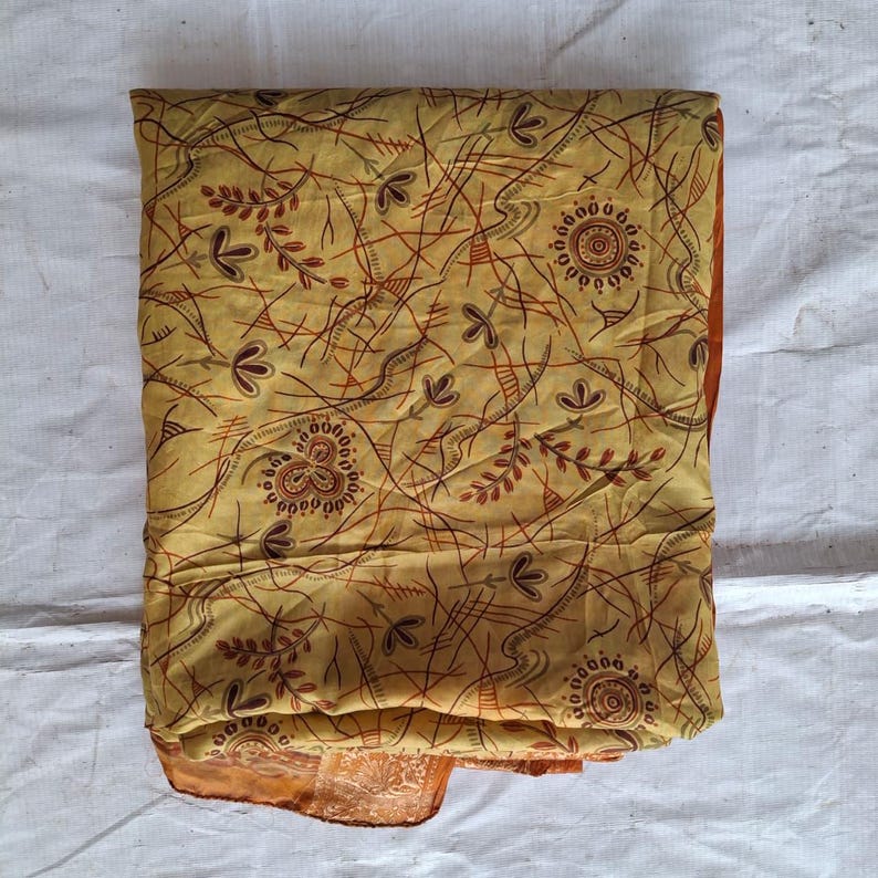 May include: A folded fabric textile featuring a mustard yellow base color with a dark brown and rust-colored pattern of thin intersecting lines, stylized floral motifs, and circular mandala-like designs printed across the entire surface.