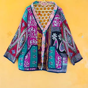 May include: A long-sleeved quilted jacket featuring a vibrant patchwork of multicoloured fabrics with intricate floral and paisley patterns. The garment includes two front pockets, a V-neckline, and visible stitching throughout the quilted construction.