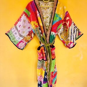 May include: A colorful patchwork kimono robe with a green tie belt. The robe features a variety of fabric patterns in red, yellow, pink, and blue. The robe is hanging on a yellow hanger against a yellow wall.