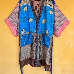 May include: A colorful, patterned kimono robe with a mix of blue, pink, and brown fabrics. The robe features a paisley design, two front pockets, and wide sleeves. It is hanging on a yellow hanger against a yellow background.