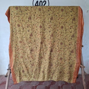 May include: A rectangular mustard yellow fabric featuring a dense, intricate pattern of dark brown swirling lines, small leaf motifs, and circular floral shapes. The fabric is framed by a wide, textured orange border with a detailed woven design.