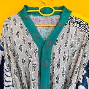 Puede incluir: Bata estampada con ribete turquesa y acento amarillo. La bata presenta un estampado de hojas blancas y negras sobre un fondo a rayas. El forro interior tiene un diseño floral. La bata está colgada de una percha amarilla.