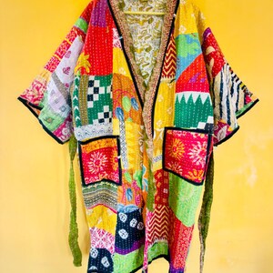 May include: A patchwork kimono-style jacket with a vibrant mix of colors and patterns. The jacket features a V-neck, two pockets, and a tie belt. The design incorporates squares of red, yellow, green, and blue fabric with various stitched details and black trim.