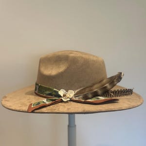 May include: A tan fedora hat with a wide brim, decorated with a patterned scarf, a white flower, and a large feather. The hat is displayed on a white stand, highlighting its features and design.