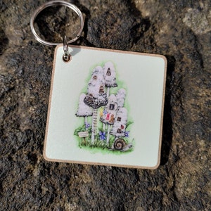 May include: Square keyring with a charming illustration of mushroom houses, a snail, flowers, and a small sign. The artwork is set against a light green background with a wooden border and a silver key ring.
