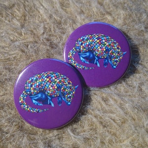 May include: Two purple round buttons with a colorful armadillo design. The armadillo is curled up with its tail tucked under its body. The armadillo is made up of many small colorful squares.