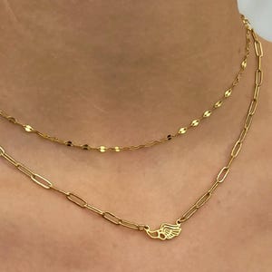 May include: Two gold necklaces layered on a person's neck. The top necklace is a delicate chain with small, flat, circular links. The bottom necklace has a paperclip chain design with a small wing charm. The necklaces are gold in color.