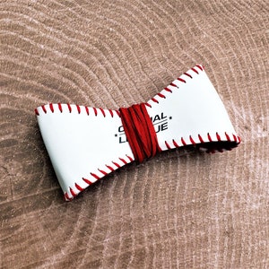 May include: White leather bow tie with red stitching and a red leather strap. The bow tie has the text "ORIGINAL LEAGUE" printed on it.