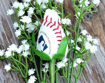 Pode incluir: Uma rosa decorativa feita de uma bola de basebol branca com costuras vermelhas, com sépalas de tecido verde e um caule verde, rodeada por flores artificiais brancas de gipsofila. A superfície da bola exibe o texto Option 10.