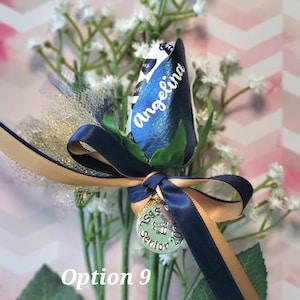 May include: A blue and white rose with the name "Angelina" on the petals, accented with navy and gold ribbons and a "Senior 2023" charm. The arrangement includes white baby's breath and green leaves. The text "Option 9" is visible.