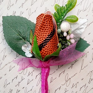 May include: A decorative boutonniere featuring a football-shaped flower bud in shades of orange and black, accented with green leaves, silver accents, and a pink ribbon. The stem is wrapped in matching pink ribbon.