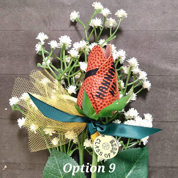 May include: A decorative flower crafted from orange basketball material with black textured dimples, featuring the name HANNAH printed in black. It is accented with gold mesh, a dark green ribbon, and a round metal charm reading CLASS OF 2025.