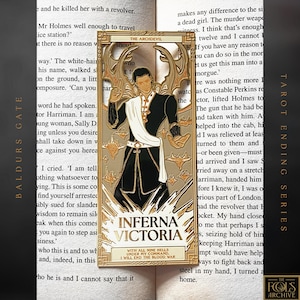 May include: A gold-coloured tarot card pin depicting a figure in black and white clothing. The card is labelled "The Archdevil" and "Inferna Victoria", with extra text. The pin is set against the backdrop of an open book.