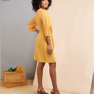 May include: A woman wearing a mustard yellow dress with a wrap design and three-quarter sleeves. The dress is paired with brown heeled sandals. The image includes the text "Simplicity Pattern S8834".