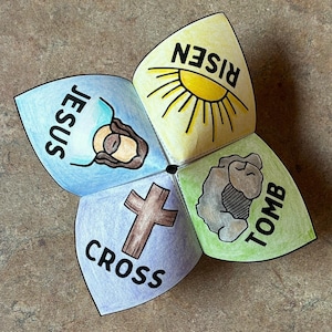 May include: A colorful paper fortune teller with the words "JESUS", "RISEN", "CROSS", and "TOMB" written on the flaps. The illustrations include a cross, a sun, and a depiction of Jesus. The fortune teller is on a brown surface.