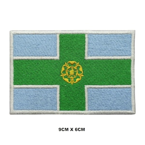 May include: An embroidered patch featuring the flag of Greater Manchester, England. The flag is a white cross on a blue background with a green cross over the top. The green cross has a gold rose in the centre. The patch is 9 cm by 6 cm.