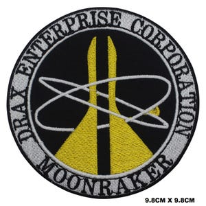 May include: A circular embroidered patch with the text "DRAX ENTERPRISE CORPORATION MOONRAKER" in white around the edge. The patch features a yellow and white design on a black background. The patch measures 9.8cm by 9.8cm.