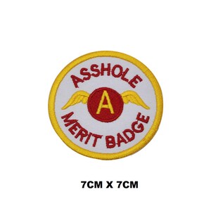 May include: A round, yellow and white embroidered patch with the text "Asshole Merit Badge" and a red "A" with wings.