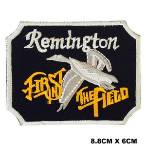 May include: A black embroidered patch with white stitching. The patch features a white goose flying with the text "Remington Fast and in the Field" in yellow and white stitching. The patch measures 8.8 cm by 6 cm.