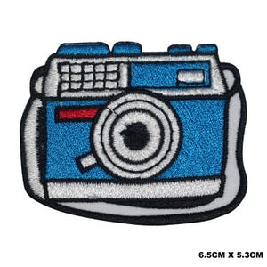 May include: An embroidered iron-on patch of a vintage camera, featuring blue and white stitching with a red stripe. The patch measures 6.5 cm by 5.3 cm.