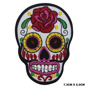 May include: Iron-on patch embroidered with a colourful sugar skull, featuring a red rose, yellow and pink eyes, and a black nose. The skull is set against a white background with green and yellow floral details. The patch measures 7.3 cm by 5.2 cm.
