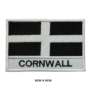 May include: Embroidered patch featuring the Cornish flag, a white cross on a black background. The word "CORNWALL" is embroidered in black on a white banner below the flag. The patch measures 3.5 inches by 2.4 inches.