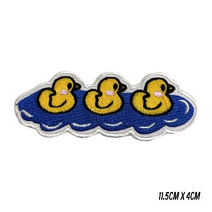 May include: An embroidered patch featuring three yellow ducks with pink cheeks swimming in a blue pond. The patch has a white border and measures 11.5 cm by 4 cm. The design shows the ducks in a row, each with a black eye and orange beak.