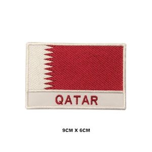 Puede incluir: Parche bordado con la bandera de Qatar. El parche es blanco con un rectángulo rojo y un triángulo blanco dentado. El texto "QATAR" está escrito en blanco debajo de la bandera. El parche mide 9 cm por 6 cm.