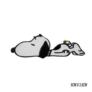 May include: Embroidered patch featuring Snoopy, the iconic cartoon dog, lying down. The patch is white with black accents and a small yellow detail. The patch measures 9cm x 3.5cm.