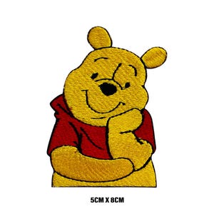 Könnte beinhalten: Gestickter Bügelaufkleber mit Winnie Puuh, einem Comic-Bären mit rotem Shirt, der lächelt und sich ans Kinn fasst. Der Aufnäher ist gelb und misst 5 cm x 8 cm.