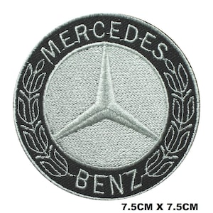 Puede incluir: Un parche bordado circular que presenta el logotipo de la estrella de tres puntas de Mercedes-Benz en color plata sobre un fondo gris claro, rodeado por un borde negro con el texto MERCEDES BENZ y un diseño de laurel. Mide 7,5 cm por 7,5 cm.