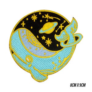 Op de afbeelding: Geborduurde patch met een lichtblauwe walvis met gele accenten, tegen een zwarte achtergrond met gele sterren en een planeet. De patch meet 8 cm x 9 cm.