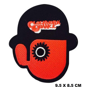May include: A black and orange embroidered patch featuring a stylized image of a person with a gear-like eye and the text "Clockwork Orange". The patch measures 9.5 cm by 8.5 cm.