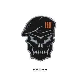 Pode incluir: Etiqueta bordada preta e cinza de uma caveira usando um boina preta com um emblema laranja. A etiqueta mede 9 cm por 7 cm.