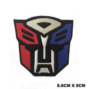 Peut inclure: Un écusson thermocollant brodé noir, rouge, blanc et bleu représentant le logo des Transformers. Le logo est un visage de robot stylisé avec un fond noir et des accents rouges et bleus. L'écusson mesure 8,8 cm sur 8 cm.