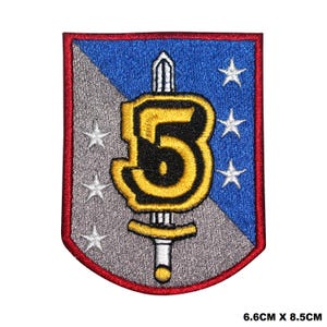 Puede incluir: Un parche bordado con forma de escudo azul y gris. El escudo contiene un número "5" dorado con contorno negro y una espada plateada en el centro. El escudo está rodeado de cinco estrellas blancas.