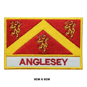 Pode incluir: Um remendo bordado com a bandeira de Anglesey, País de Gales. A bandeira é vermelha com um galão amarelo e três leões amarelos. O remendo mede 9 cm por 6 cm e tem uma borda branca com a palavra "Anglesey" em letras amarelas.