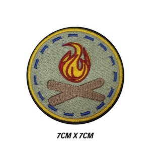 Op de afbeelding: Een ronde patch met een kampvuurontwerp. De patch heeft een lichtgroene achtergrond, een gele rand en blauwe stiksels. Het kampvuur heeft een rood en gele vlam boven twee bruine gekruiste boomstammen. Afmetingen: 7 cm x 7 cm.