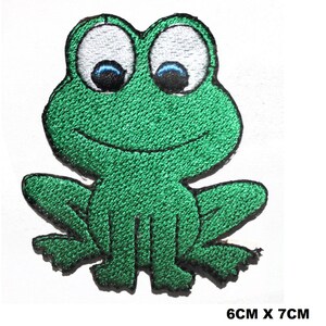 Può includere: Toppa ricamata verde da stirare con un disegno di rana dei cartoni animati con occhi bianchi e pupille blu. La rana è seduta con le gambe incrociate e ha un ampio sorriso. La toppa misura 6 cm per 7 cm.